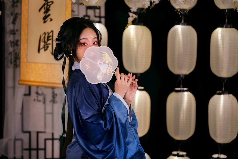 May include: A woman in a blue robe holds a white fan with a rabbit design in front of her face. The fan has a gold handle. The background is a wall with white lanterns.