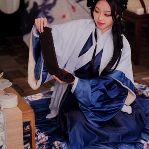 May include: A woman wearing a blue and white robe sits on a patterned rug, holding a rolled up dark brown fabric. The robe has a white fur trim. The woman has long black hair and is looking down at the fabric.
