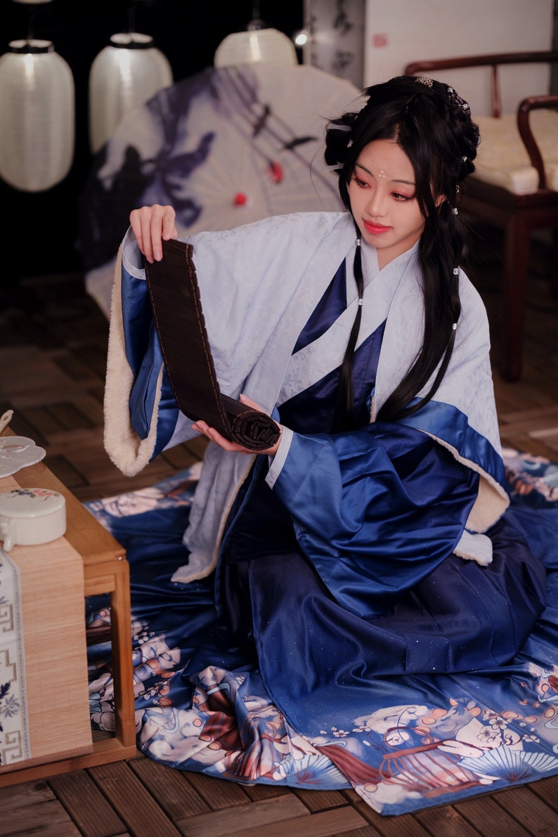 May include: A woman wearing a blue and white robe sits on a patterned rug, holding a rolled up dark brown fabric. The robe has a white fur trim. The woman has long black hair and is looking down at the fabric.
