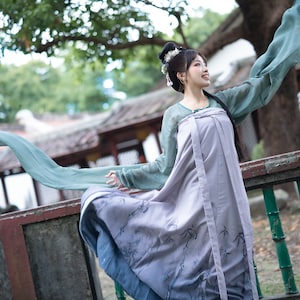 Puede incluir: Una mujer con un vestido tradicional chino con una manga larga y fluida. El vestido es de color azul claro y morado con un estampado floral. La mujer sonríe y sostiene la manga de su vestido en su mano. Está de pie frente a una valla de madera con bambú verde.