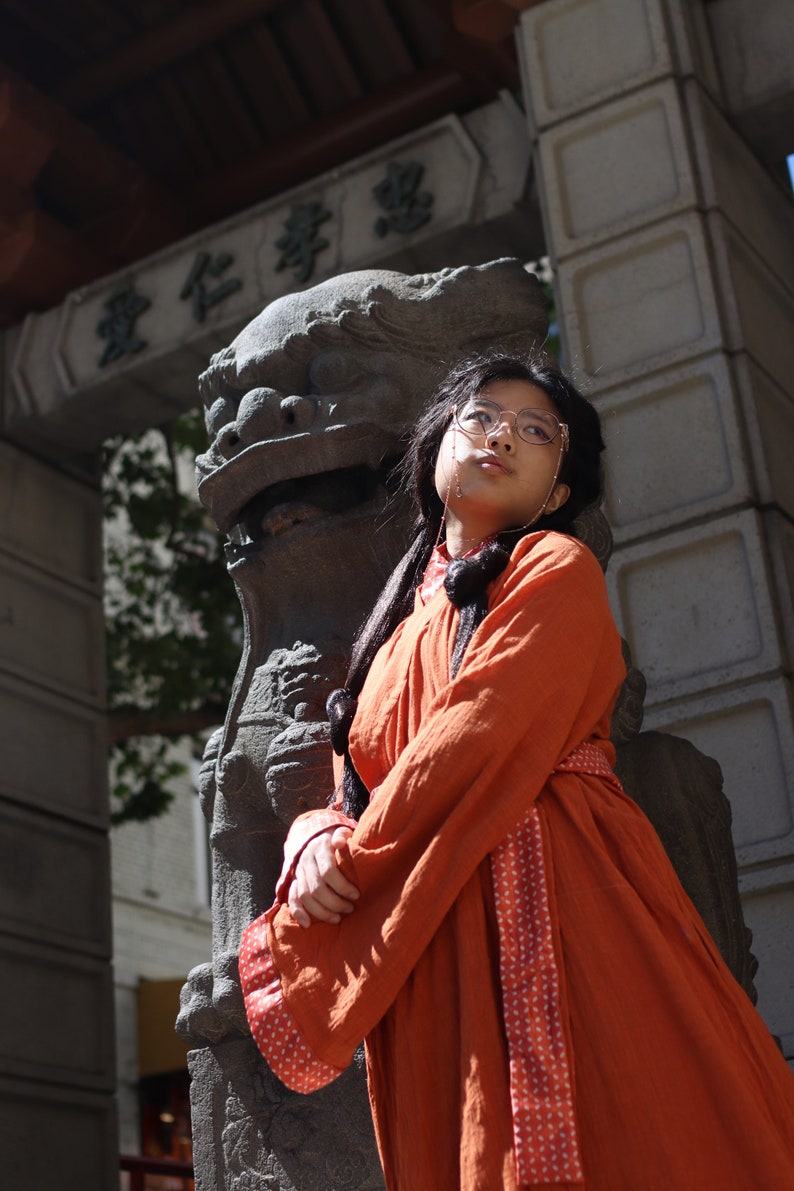 May include: A person wearing a long, orange robe with a red and white patterned sash stands in front of a large, grey stone lion statue. The statue is in front of a building with a sign that reads "愛仁學校".