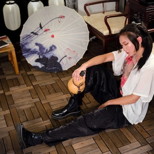 Puede incluir: Una mujer con una camisa blanca y pantalones negros se sienta en un suelo de madera. Ella sostiene una botella con forma de calabaza en su mano. El suelo está hecho de baldosas de madera. Hay una sombrilla blanca y azul en el fondo.