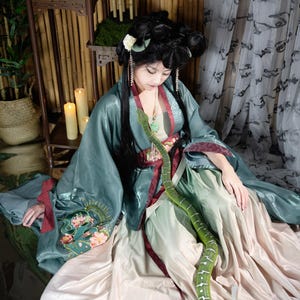 Puede incluir: Una persona con un vestido tradicional asiático verde azulado y rojo con bordados florales. Una serpiente verde brillante con marcas blancas está sobre el regazo de la persona. El fondo incluye bambú y velas.