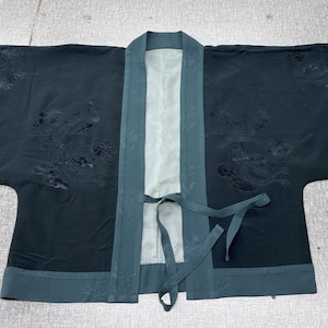 Puede incluir: Una bata estilo kimono de color verde azulado oscuro con forro y ribete verde azulado claro. La bata tiene un cierre de lazo y presenta un patrón sutil en relieve. Las mangas son anchas.