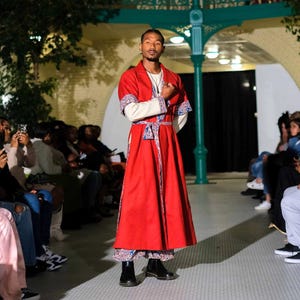 Puede incluir: Una persona lleva una túnica roja con un ribete estampado en azul y blanco. La túnica tiene una faja blanca y se lleva sobre una camisa blanca de manga larga. La persona está de pie en un escenario con una audiencia en el fondo.
