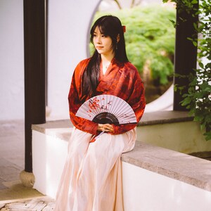 May include: A woman in a red and white kimono sits on a white wall with a hand fan. She is holding the fan in front of her and looking to the side. The background is a garden with a circular window.