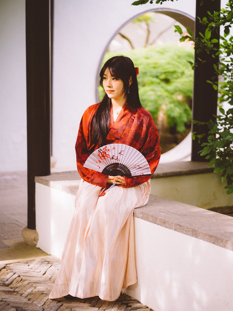 May include: A woman in a red and white kimono sits on a white wall with a hand fan. She is holding the fan in front of her and looking to the side. The background is a garden with a circular window.