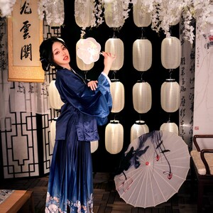 May include: A woman in a blue and white patterned dress holds a white fan with a floral design. She is standing in front of a wall with white paper lanterns and a scroll with Chinese characters that read "心香一瓣".
