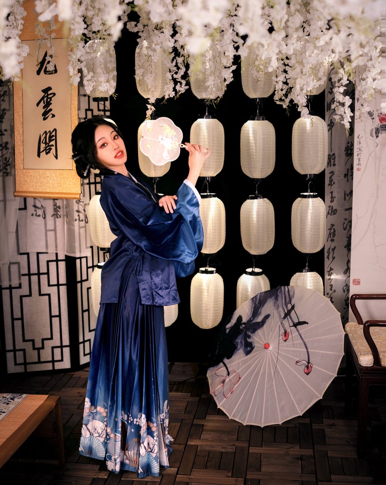May include: A woman in a blue and white patterned dress holds a white fan with a floral design. She is standing in front of a wall with white paper lanterns and a scroll with Chinese characters that read "心香一瓣".