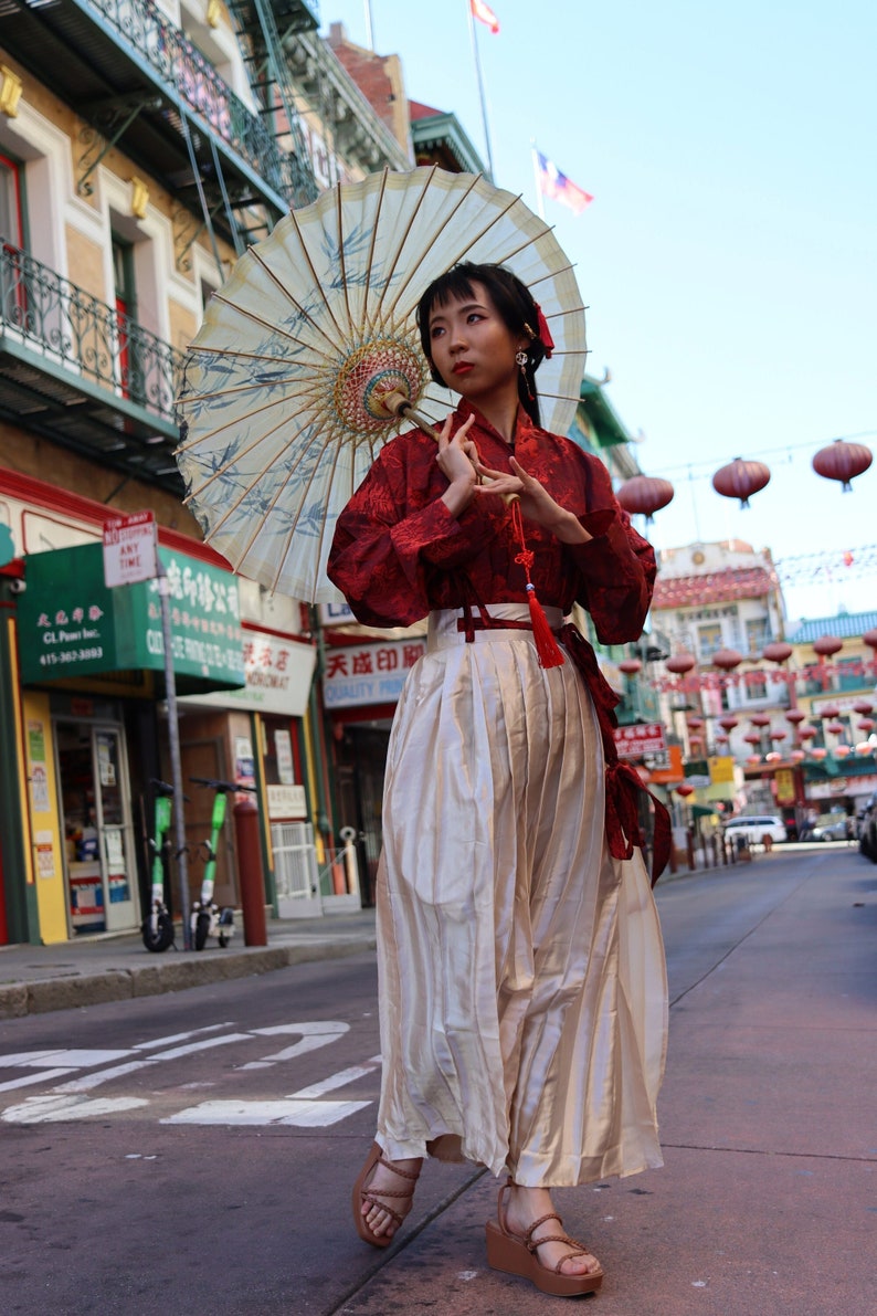 May include: A woman wearing a red and white traditional Chinese outfit with a white skirt and a red top. She is holding a white and gold umbrella and is standing on a street with red lanterns hanging overhead.