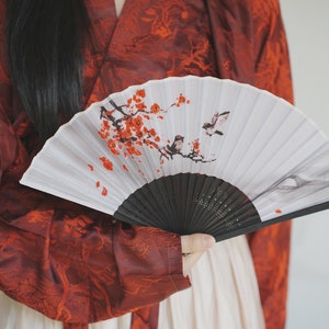 May include: A person wearing a red and white patterned robe holds a white folding fan with a black handle. The fan has a painted design of a cherry blossom branch with red flowers and two birds.