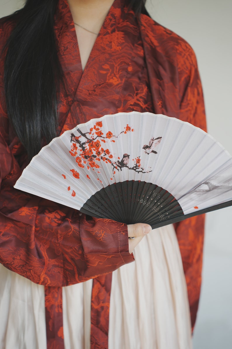 May include: A person wearing a red and white patterned robe holds a white folding fan with a black handle. The fan has a painted design of a cherry blossom branch with red flowers and two birds.
