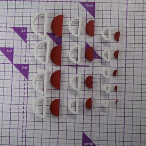 Op de afbeelding: Een verzameling witte en rode halve cirkel koekjessnijders van verschillende formaten. De snijders zijn gerangschikt op een paarse snijmat met raster. De snijders zijn gemaakt van plastic en worden waarschijnlijk gebruikt om te bakken of te knutselen.