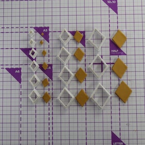 Op de afbeelding: Een set witte plastic diamantvormige koekjesuitstekers in verschillende maten, met gouden fondant uitsnijdingen in dezelfde vormen.