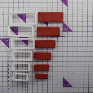 May include: A set of six white plastic rectangle-shaped cookie cutters with red fondant cutouts of the same shape. The cutters are arranged in descending size order from top to bottom. The fondant cutouts are also arranged in descending size order from top to bottom. The background is a purple grid.