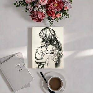 May include: A book titled P.J. Lenda Dług wdzięczności with a black and white pencil sketch of a person holding a child on the cover. Beside it lies a light gray notebook labeled COUNTY, a white cup of coffee with a black spoon, and a floral arrangement.