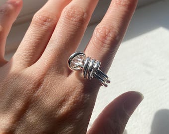 May include: A silver-colored ring with a unique knot design. The ring features a series of intertwined bands, creating a textured and eye-catching appearance. The ring is worn on a finger, showcasing its intricate details and reflective surface.