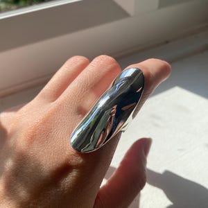 May include: A large, polished silver ring with a smooth, elongated design. The ring reflects the surrounding light, creating a bright, shiny appearance. The ring is worn on a finger, showcasing its size and shape. A modern piece of jewelry.