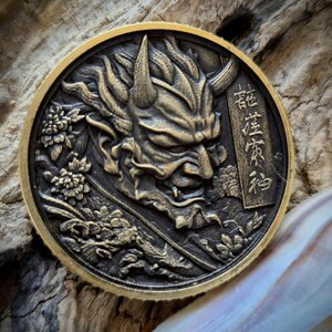 May include: A circular bronze-toned metal coin featuring a high-relief embossed design of a horned demon mask. The artwork includes intricate floral patterns and a vertical rectangular panel with Japanese kanji characters on the right side.