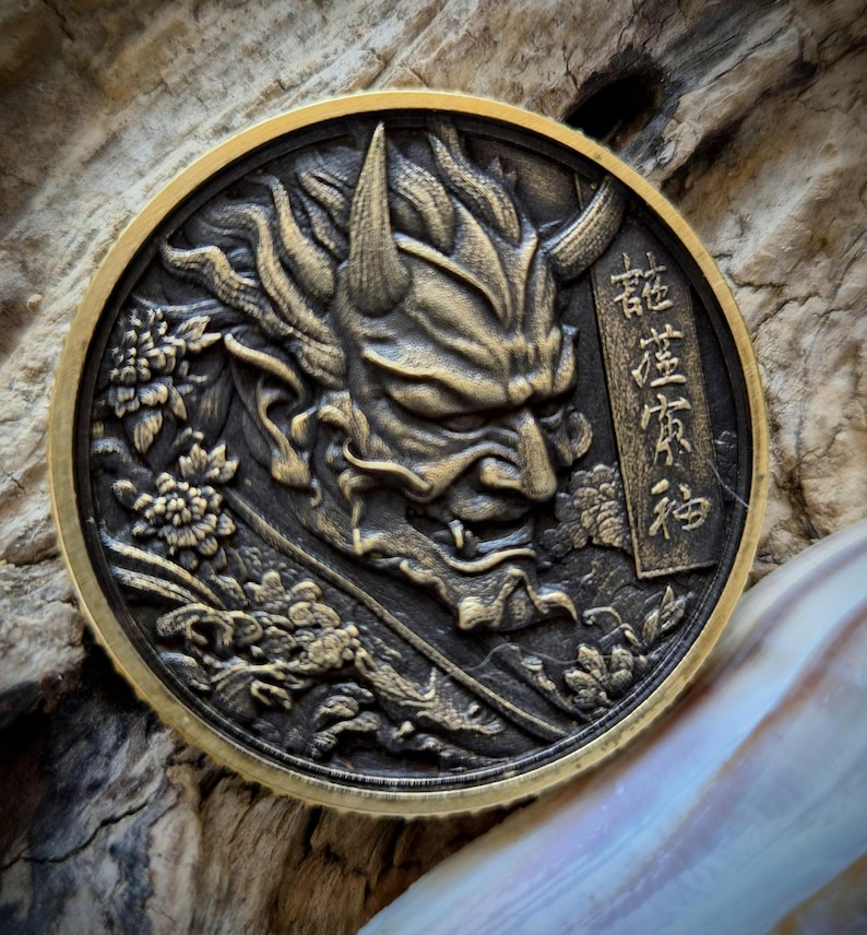 May include: A circular bronze-toned metal coin featuring a high-relief embossed design of a horned demon mask. The artwork includes intricate floral patterns and a vertical rectangular panel with Japanese kanji characters on the right side.