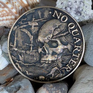 Puede incluir: Una moneda de color bronce con un diseño de calavera y barco pirata. La moneda tiene las palabras "NO QUARTER" alrededor del borde. La moneda descansa sobre un lecho de rocas, conchas y arena.