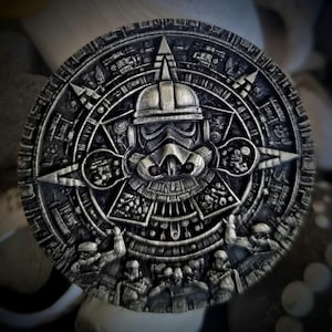 Può includere: Scultura circolare in rilievo metallico, con un elmetto da Stormtrooper al centro, circondato da intricati disegni di ispirazione azteca. La scultura ha un aspetto invecchiato e antico, con un colore grigio scuro e dettagli in rilievo. Il tema è una fusione di Star Wars e arte antica, diametro circa 10 cm.