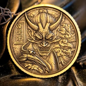 May include: A circular bronze-toned medallion featuring a relief carving of a horned demon mask with sharp teeth and a topknot hairstyle. The design includes intricate swirling patterns and a rectangular frame containing Japanese kanji characters.