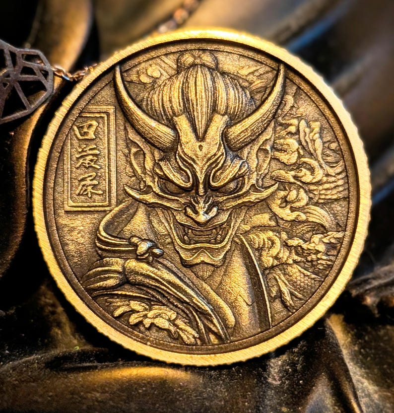 May include: A circular bronze-toned medallion featuring a relief carving of a horned demon mask with sharp teeth and a topknot hairstyle. The design includes intricate swirling patterns and a rectangular frame containing Japanese kanji characters.