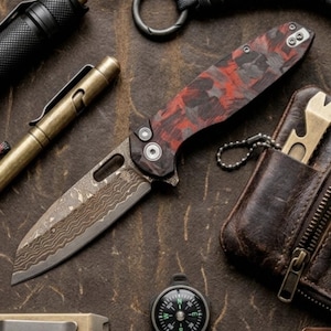 May include: A folding pocket knife with a red and black marbled handle and a patterned Damascus steel blade, accompanied by a brass pen, a small round compass, a black flashlight, a metal pry bar, and a brown leather wallet with a metal tool.