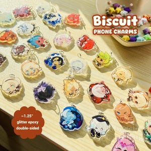 May include: A selection of double-sided glitter epoxy phone charms, each with a unique character design. The charms are approximately 3.18 cm in size and come in various colours and styles. The image also includes the text "Biscuit PHONE CHARMS".