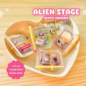 Peut inclure: Plusieurs Alien Stage Bento Charms dans des boîtes en plastique transparent, maintenus par des élastiques jaunes, présentés dans un plat en forme de cœur. Chaque breloque mesure environ 5,4 cm et est recto-verso. Le texte indique "Alien Stage Bento Charms."