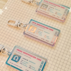 May include: Three ID card keychains with iridescent edges. Each card has a different Greek letter: Alpha, Beta, and Omega. The cards have a clear plastic overlay and a silver clasp. The cards have text including "Identification Card" and other details.