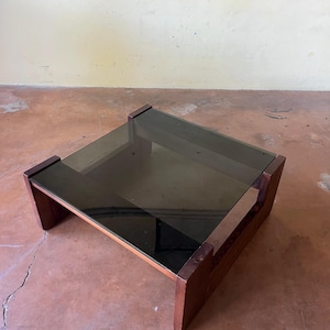 May include: A low, rectangular coffee table with a dark, tinted glass top. The table has a dark wood frame and legs. The glass top is approximately 36 inches long and 36 inches wide.