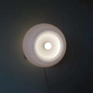 Può includere: Un'applique da parete bianca e circolare con una lampadina centrale luminosa. L'apparecchio ha una finitura liscia e lucida ed è montato su una parete grigio scuro. Un cavo bianco si estende dalla luce.