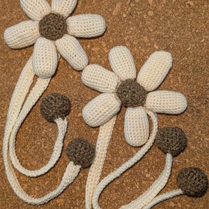 May include: Two crocheted white daisy flowers with brown centers, each attached to a long white crocheted cord with brown crocheted balls at the ends.