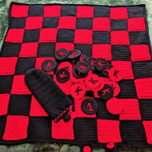 May include: A handmade crochet checkers set featuring a red and black checkerboard blanket. The set includes red and black checkers pieces, some marked with a "K", and a black storage bag. The blanket measures approximately 30 inches square.