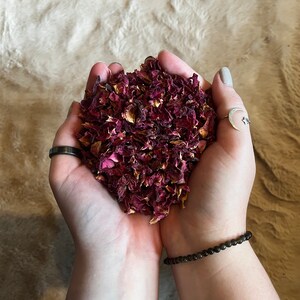 May include: A handful of dried rose petals, a deep red colour, held in two hands. The petals are loose and scattered, creating a textured look.