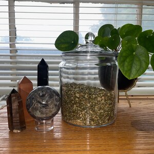 May include: A glass jar with a lid filled with dried herbs, a black and white marble sphere on a clear stand, a brown crystal, and a black crystal on a wooden surface.