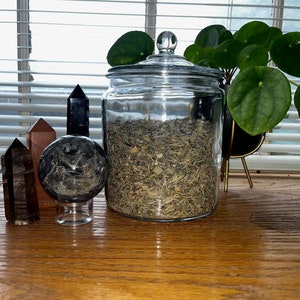 May include: A glass jar filled with dried herbs, sitting on a wooden table with two small, dark-coloured stones and a brown stone. A green plant with round leaves is in the background.