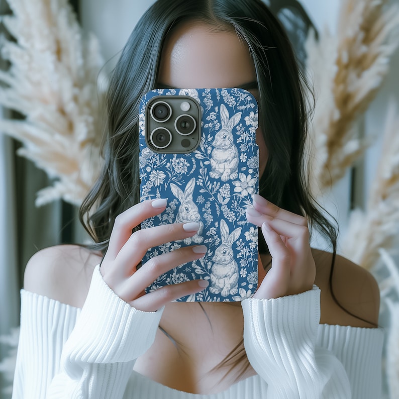 May include: A blue phone case with a white floral and bunny pattern. The case has a camera lens cutout and a white bunny with a flower in its mouth.