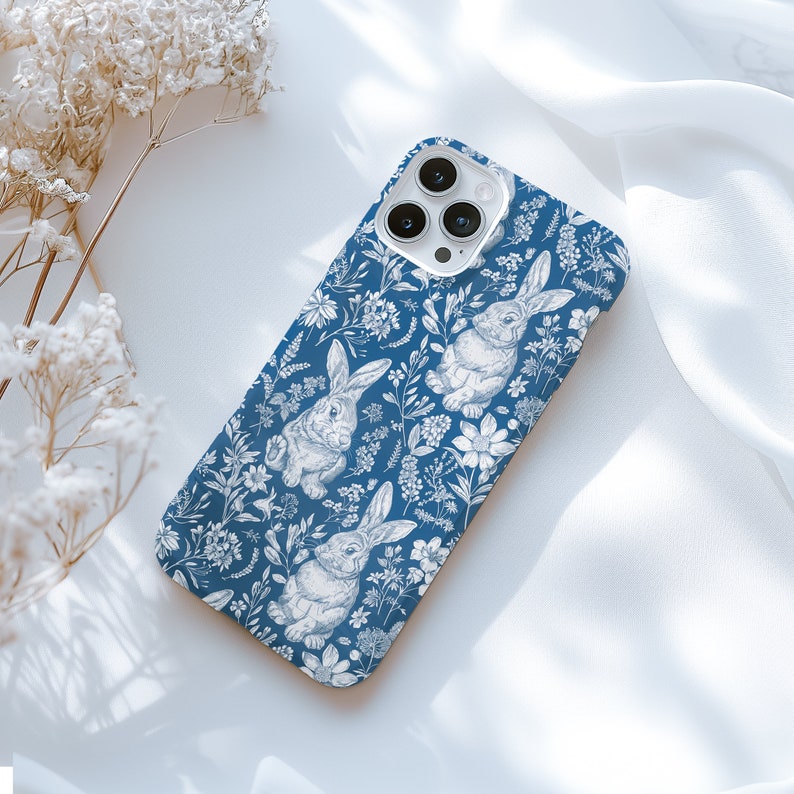 May include: A blue phone case with a white floral pattern and bunnies. The case has a camera lens cutout.