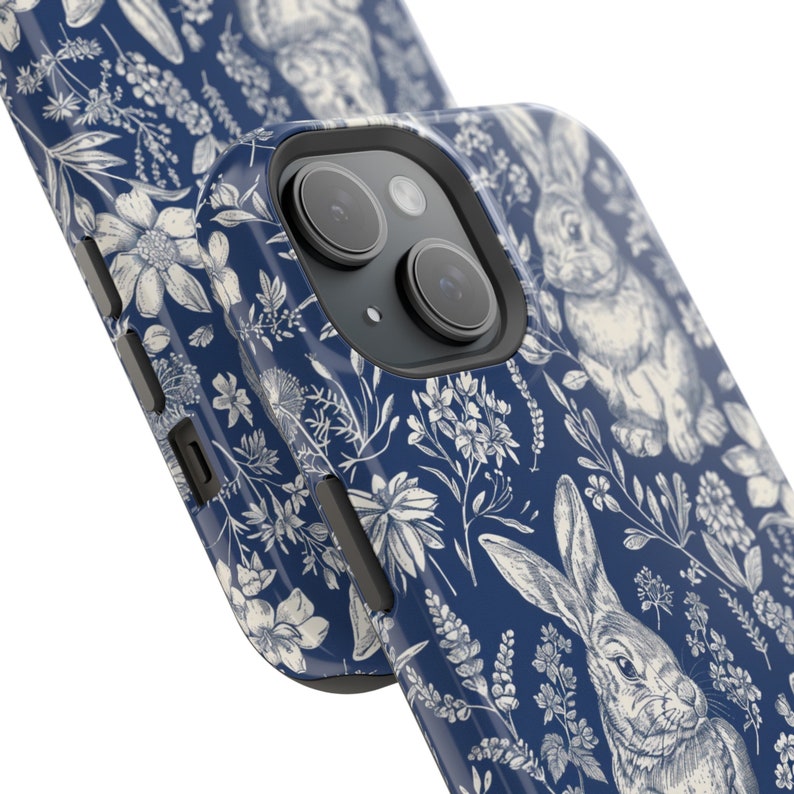 May include: A blue phone case with a white floral and bunny pattern. The case has a raised camera bump and a cutout for the phone's buttons.
