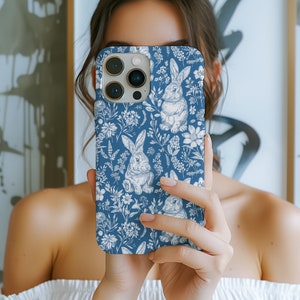 May include: A blue phone case with a white floral pattern and bunnies. The case has a camera lens cutout and a white outline.