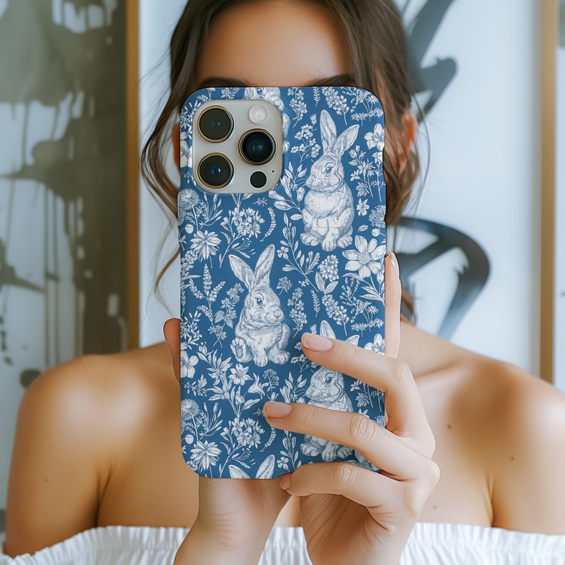 May include: A blue phone case with a white floral pattern and bunnies. The case has a camera lens cutout and a white outline.
