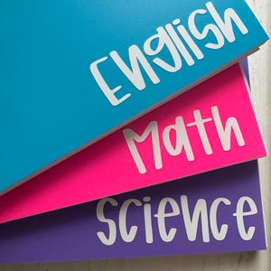 May include: Three notebooks in blue, pink, and purple, labeled "English," "Math," and "Science" in white, handwritten-style lettering. The notebooks are stacked, with the top one blue, the middle one pink, and the bottom one purple.