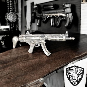 May include: A silver-colored metal replica of a submachine gun. The gun is on a dark wood surface.