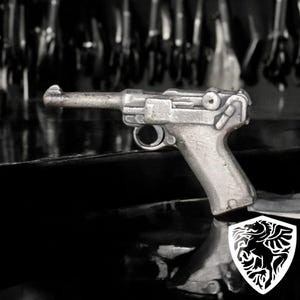 May include: A silver-colored replica of a pistol, with a detailed design and a textured surface. The pistol is sitting on a black surface.