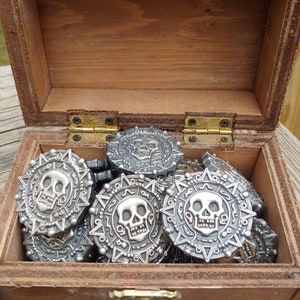 May include: A wooden treasure chest filled with silver-colored coins. Each coin features a skull design in the center, surrounded by an intricate pattern. The chest is open, revealing the coins inside.