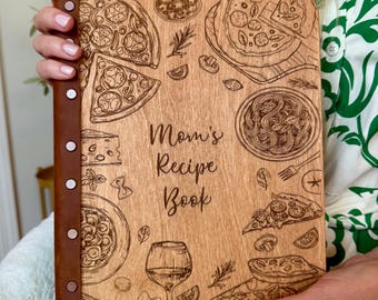 Puede incluir: Libro de recetas de madera con lomo de cuero marrón. La portada está grabada con ilustraciones de pizza, pasta y una copa de vino. El texto "Mom's Recipe Book" está centrado en la portada.