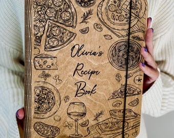 May include: A rectangular wooden recipe book with a light brown finish, featuring laser-engraved illustrations of pizza, pasta, cheese, and wine. The center text reads Olivia's Recipe Book in a script font. A thin black elastic band secures the cover.